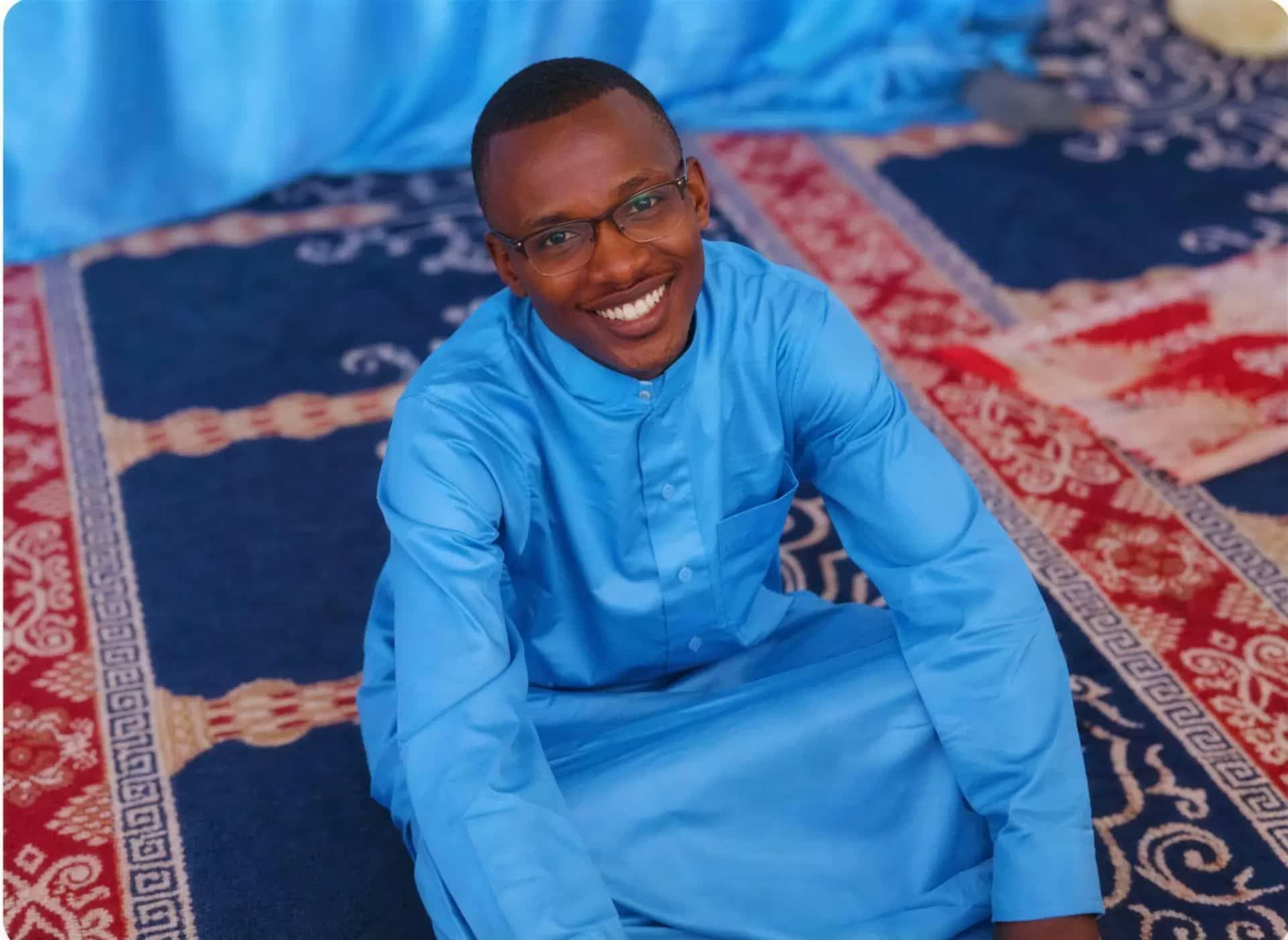 A man in a light blue tunic kneeling on a patterned prayer rug at a community centre in Kenya